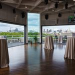 A modern event space with high ceilings and city views, featuring cocktail tables with white tablecloths, large windows, and an outdoor terrace overlooking a skyline with trees and tall buildings.