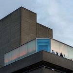 A group of people stand on a balcony outside a modern concrete building at sunset, with light reflecting off frosted glass panels and a cloudy sky overhead.