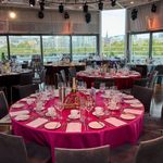 A modern banqueting hall with large windows and city views, featuring round tables covered in magenta tablecloths, set with glassware, plates, menus, and decorative centrepieces, ready for an elegant event.