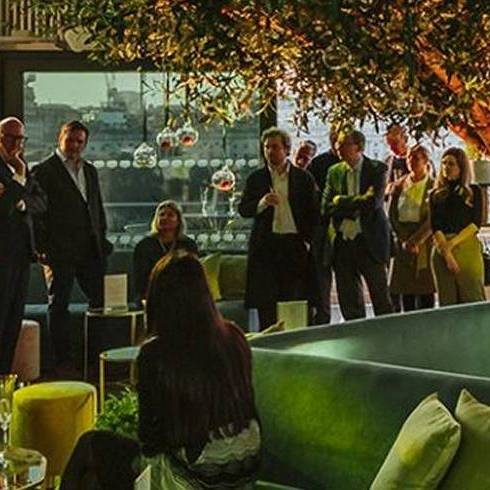 People in formal attire gather around a large, illuminated indoor tree at a modern event space with city views, comfortable seating, and plants, creating a stylish, vibrant atmosphere.