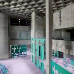 A large, modern interior with concrete walls and pillars, lilac carpeting, and green partitions with circular cut-outs. Rows of light purple folding chairs line the walls, creating an open, geometric space.