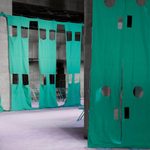 Large teal fabric panels with cut-out shapes hang from tall concrete pillars in an industrial indoor space with a purple floor. Metal chairs are visible in the background.