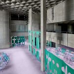 A spacious, modern interior with concrete walls and ceiling, purple carpet, mint green folding chairs, and green fabric partitions decorated with abstract cut-out shapes. The space feels open and geometric.