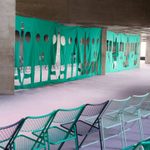 Rows of empty wire chairs in teal and white face a large green fabric partition with various-sized cut-out shapes, set in a spacious, modern concrete interior.