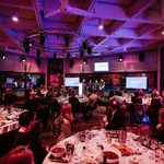 A large group of people in formal attire sit at round tables for a dinner event, watching speakers on a stage in a modern, purple-lit venue with city views visible through the windows.