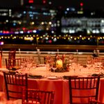 Elegant round tables set for dinner with glassware, cutlery, serviettes, and a candle centrepiece, on an outdoor terrace at night with city lights and fairy lights in the blurred background.