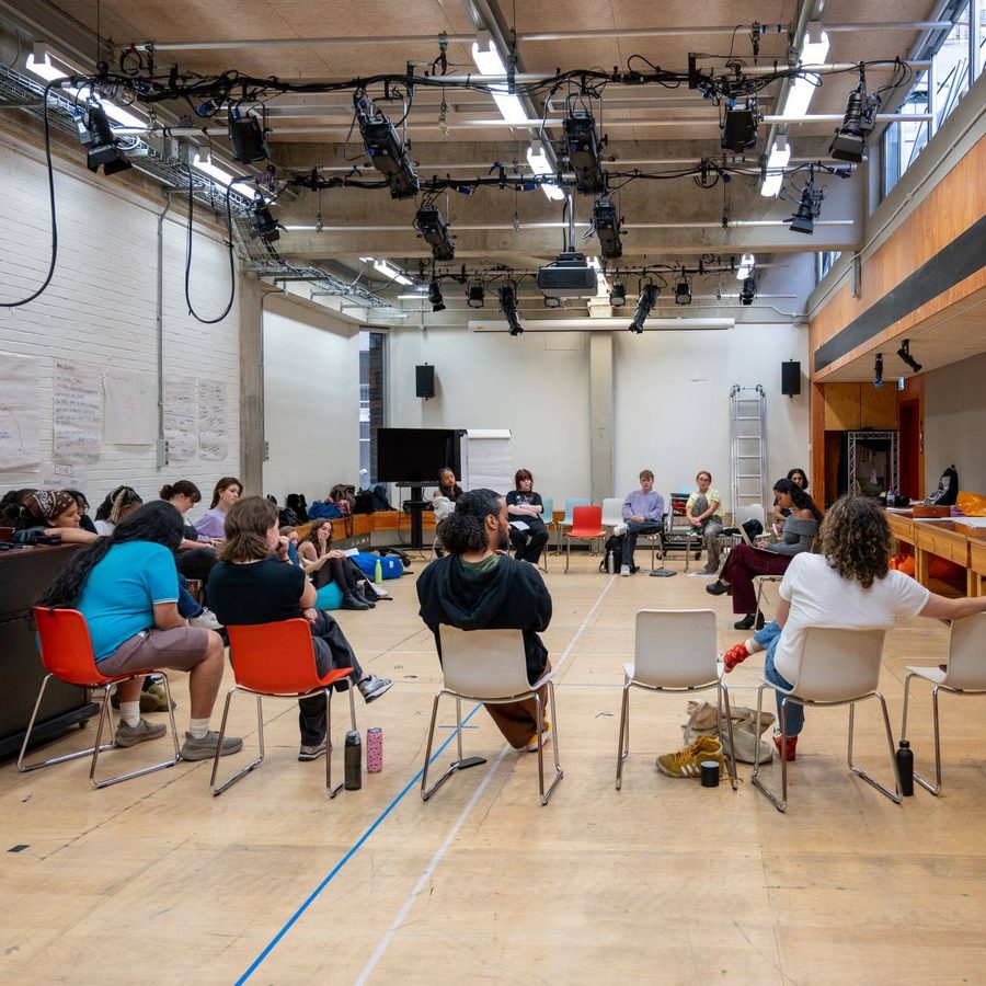A group of people sit in a large circle of chairs in a spacious room with high ceilings, a piano, and white brick walls, engaged in discussion or a workshop.