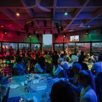 A large group of people sit at round tables in a dimly lit event space with colourful lights, listening to a speaker on stage. Large windows reveal a cityscape outside.