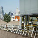 A modern outdoor terrace with yellow chairs and potted plants overlooks a cityscape of tall skyscrapers and historic buildings on a clear day.