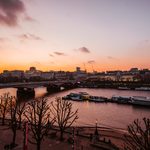 A sunset view over the River Thames in London, with silhouetted leafless trees, boats moored along the water, and buildings and bridges against a colourful orange and purple sky.
