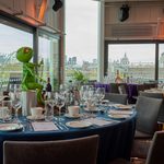 A dining table set for an event with glasses, plates, and menus, featuring a Kermit the Frog puppet centrepiece by the window. City buildings and trees are visible outside through large glass windows.