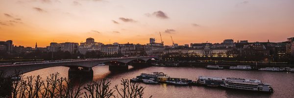 nt-events-our-spaces-scaled-aspect-ratio-1800-600 A wide view of a city river at sunset, with a bridge crossing over the water, boats moored along the riverbanks, and buildings silhouetted against an orange and purple sky. Leafless trees line the riverside.