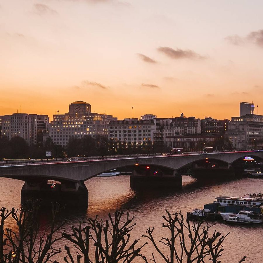 A wide view of a city river at sunset, with a bridge crossing over the water, boats moored along the riverbanks, and buildings silhouetted against an orange and purple sky. Leafless trees line the riverside.