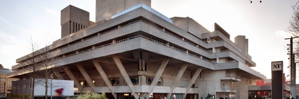 A large, angular concrete building with multiple levels and sharp lines, supported by thick pillars. The modernist structure is surrounded by cityscape elements and people walking nearby.