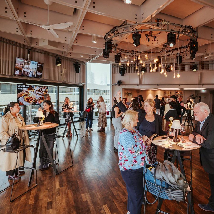 RyanDinham_160925_NTdeck_203_small-aspect-ratio-1800-600 A group of people socialise and converse around tall tables in a modern, warmly lit event space with wooden floors and hanging lights. Some stand whilst others sit, and food and drinks are visible on the tables.
