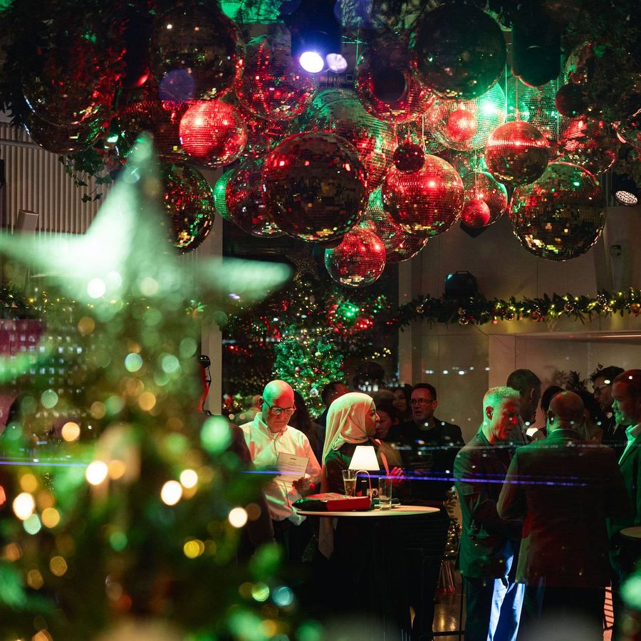 People gather at a festive indoor event decorated with large red and gold baubles, twinkling fairy lights, and garlands, creating a warm, holiday atmosphere. Some guests stand and talk around tables.