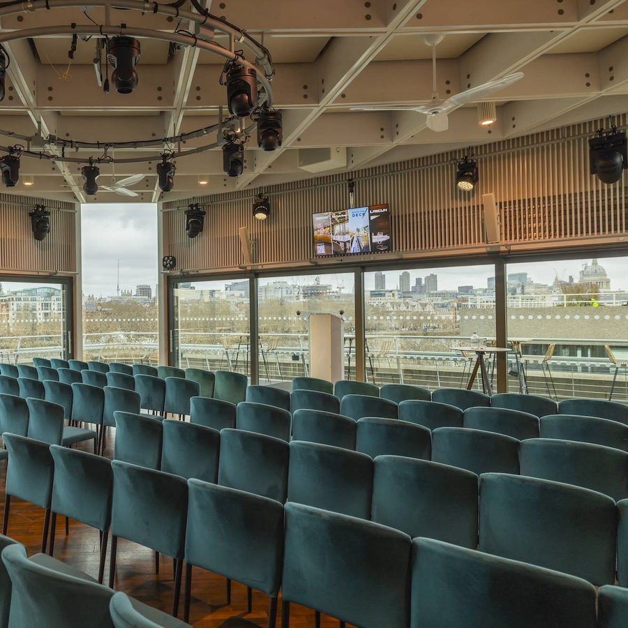 Rows of teal chairs are arranged in an empty, modern conference room with large windows overlooking a cityscape. Ceiling-mounted stage lights and a wall-mounted screen are visible. Daylight streams in through the windows.