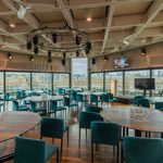 Spacious function room with large windows overlooking a cityscape, round wooden tables with blue velvet chairs, ceiling lights, and audiovisual equipment. Glasses and water jugs are on some tables.