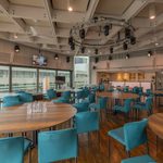 A spacious, modern event room with round wooden tables, teal upholstered chairs, wooden floors, large windows, and ceiling lights. Glass water bottles and glasses are set on the tables. The room is well-lit and airy.