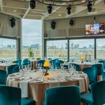 A modern dining room with round tables set for an event, featuring teal chairs, white tablecloths, and large windows overlooking a city skyline on a cloudy day.