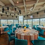 Elegant dining room with round tables set for an event, teal chairs, large windows offering a cityscape view, and ceiling lights arranged in a circular structure overhead.